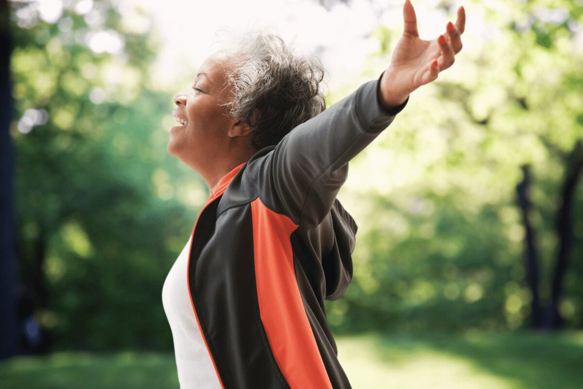 A woman standing outdoors with her arms open, eyes closed, and face lifted toward the sunlight, appearing calm, free, and connected.