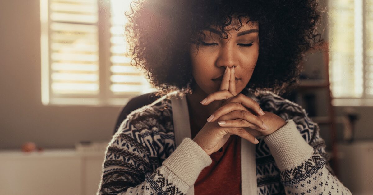 A woman sitting indoors with her eyes closed and hands gently resting together, appearing calm and focused in meditation.