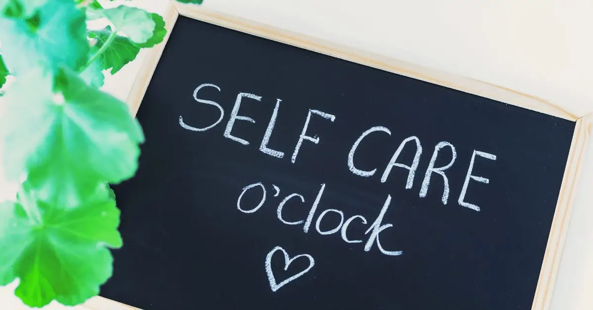 Small chalkboard sign reading ‘Self care o’clock’ with a heart, next to a green plant on a light background.