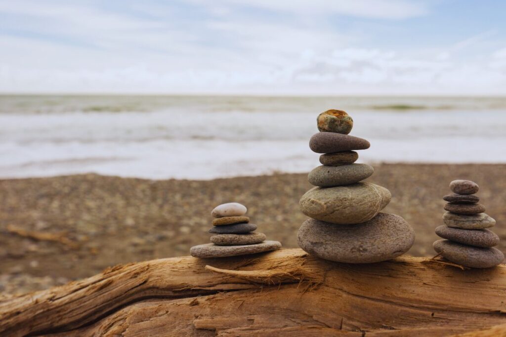 Balanced stack of smooth stones on driftwood near the ocean, representing stability, consistency, and steady personal growth.