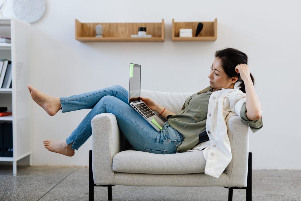 A woman reclining comfortably in an armchair while using a laptop, appearing relaxed and disengaged in a quiet living space.