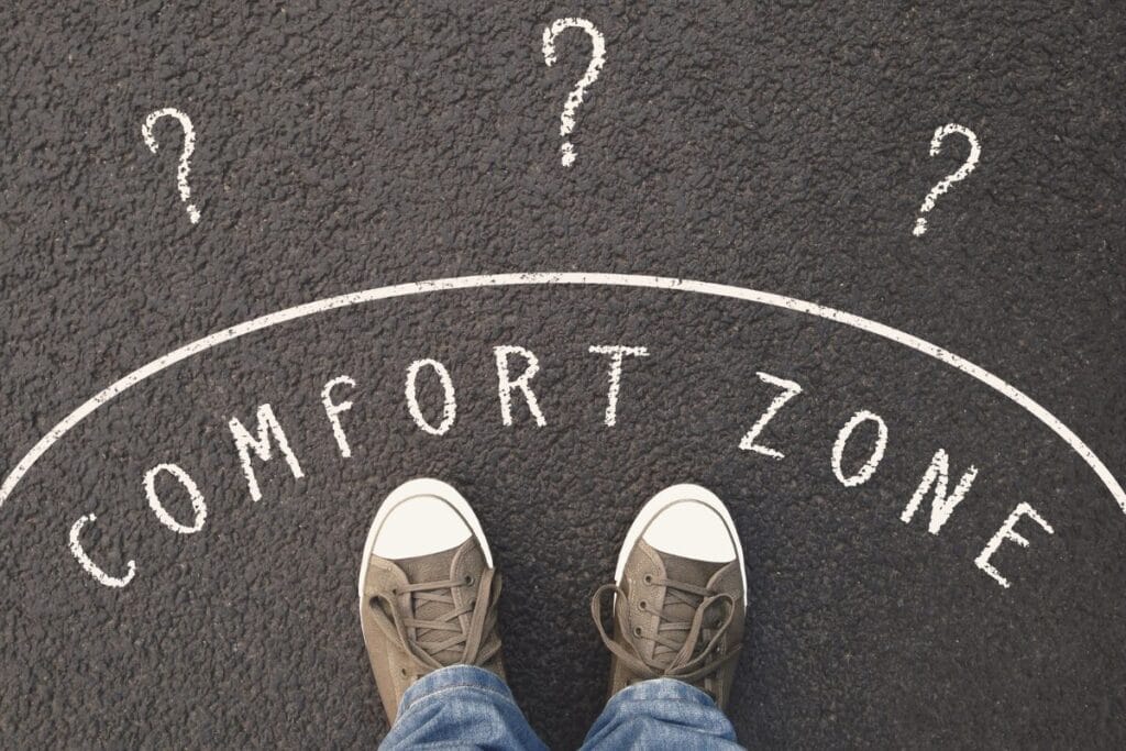 A pair of shoes standing on pavement marked with the words “comfort zone” and several question marks drawn ahead, illustrating a moment of uncertainty or decision.