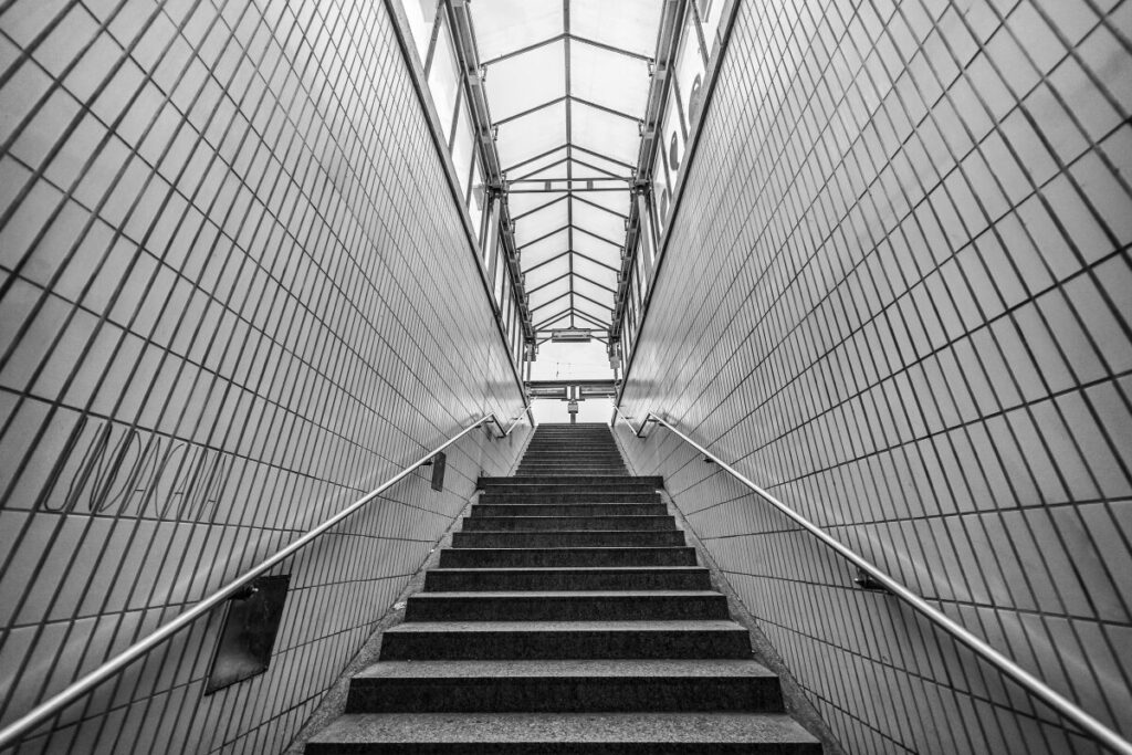 A black-and-white image of a stairway leading upward through a tiled passage with handrails on both sides and light visible at the top.