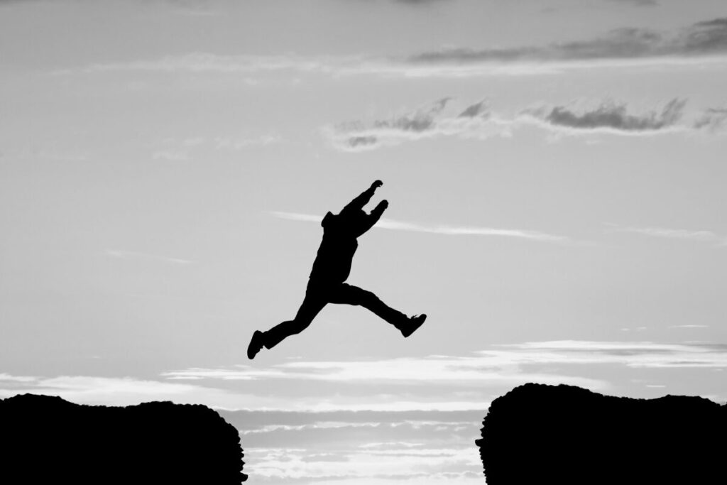 A silhouetted person midair, jumping across a gap between two elevated surfaces against a cloudy sky.