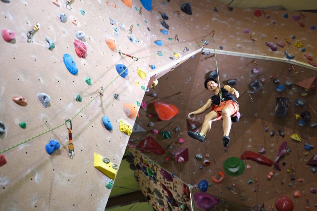 Person rock climbing indoors, smiling while suspended on a rope against a colorful climbing wall.
