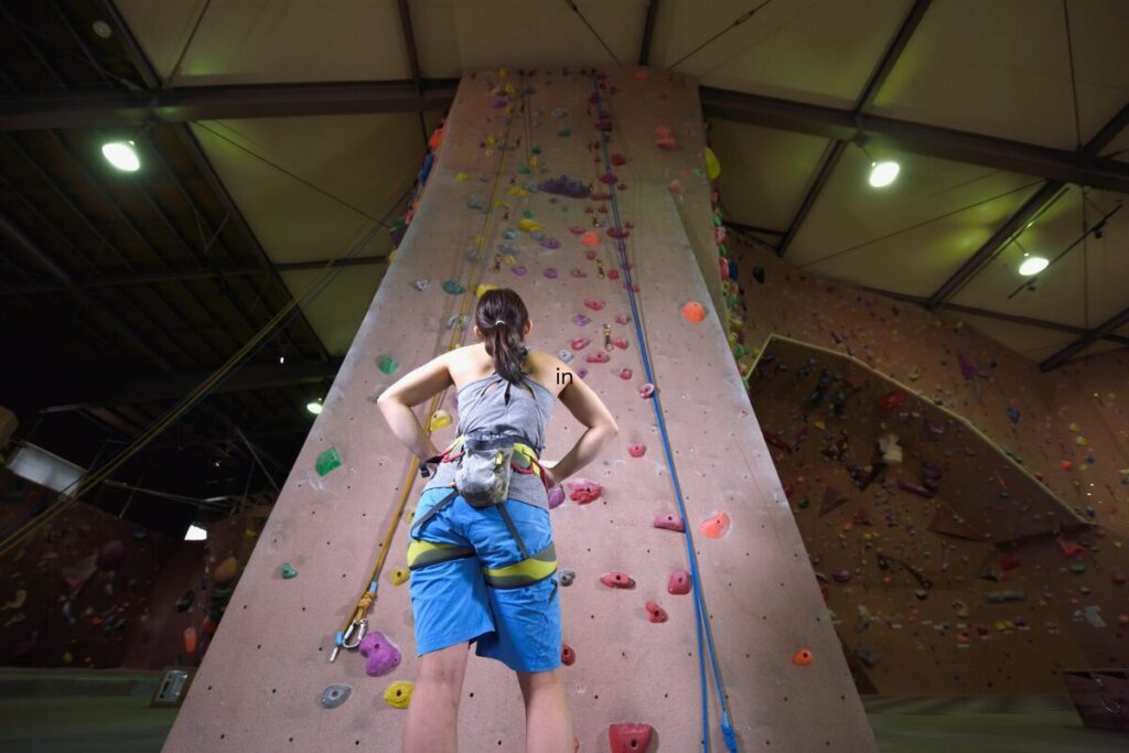Woman standing at the base of an indoor climbing wall, looking upward and preparing to climb.