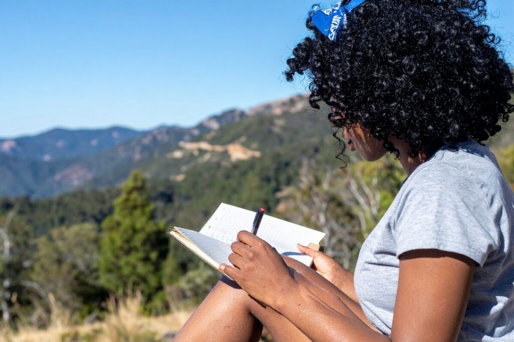 Woman sitting outdoors journaling in a notebook with mountains in the background, illustrating how starting small builds momentum