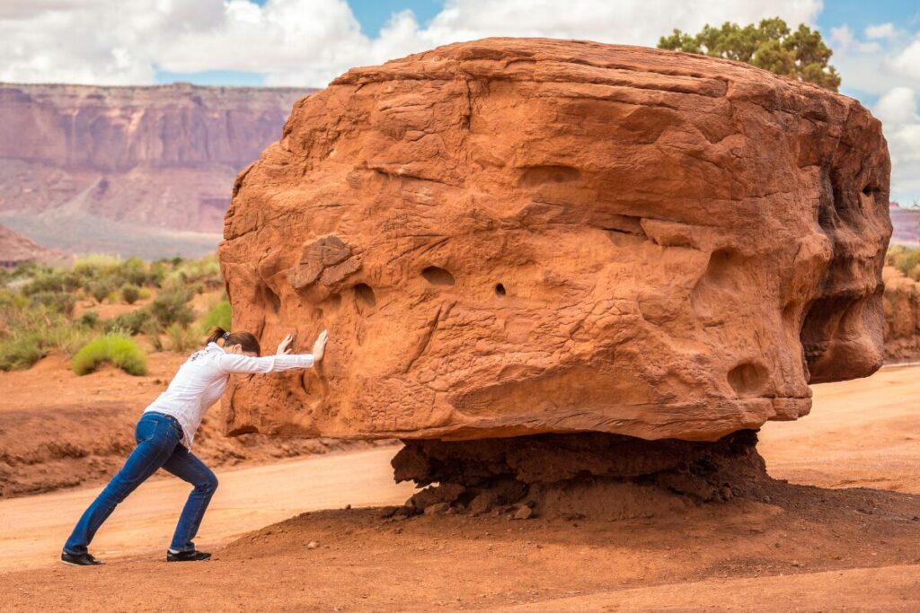 Woman pushing against a large rock in a desert landscape, representing effort and challenge.