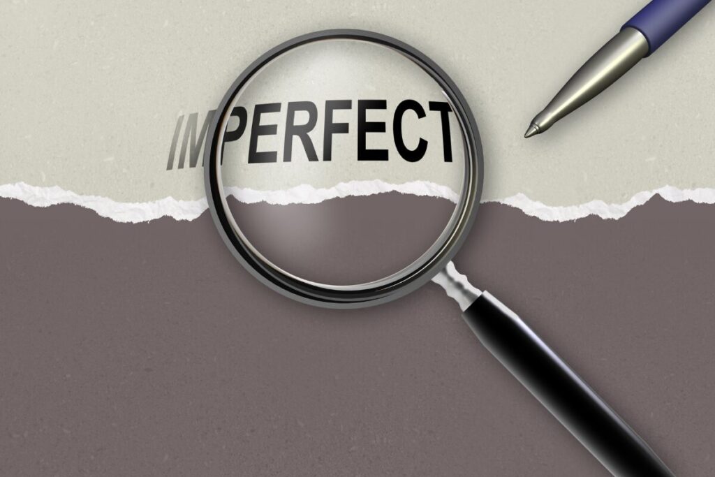Magnifying glass placed over the word “imperfect” on torn paper, representing how to stop being a perfectionist and embrace imperfect progress.