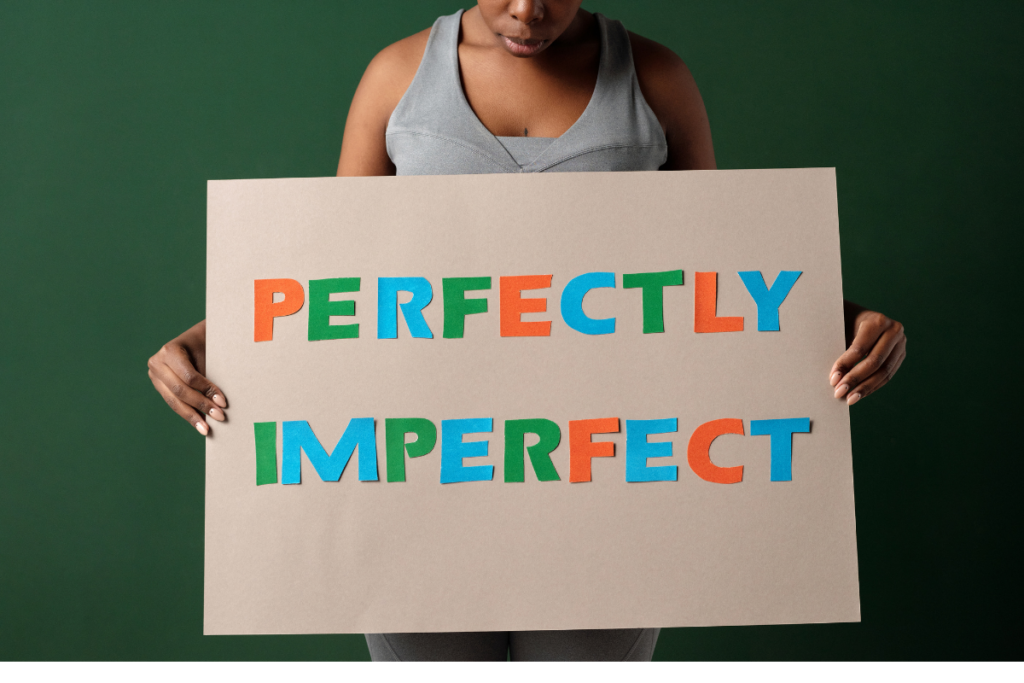 Person holding a large sign that reads “Perfectly Imperfect” in colorful letters against a green background.