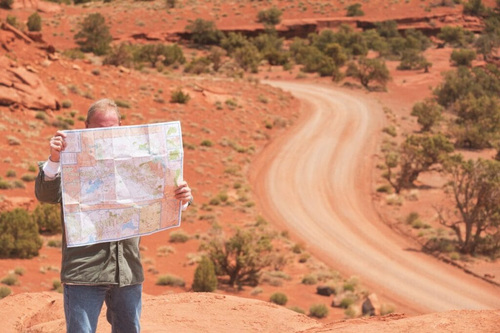 Man standing in a red desert landscape holding a large map while a winding dirt road stretches into the distance.