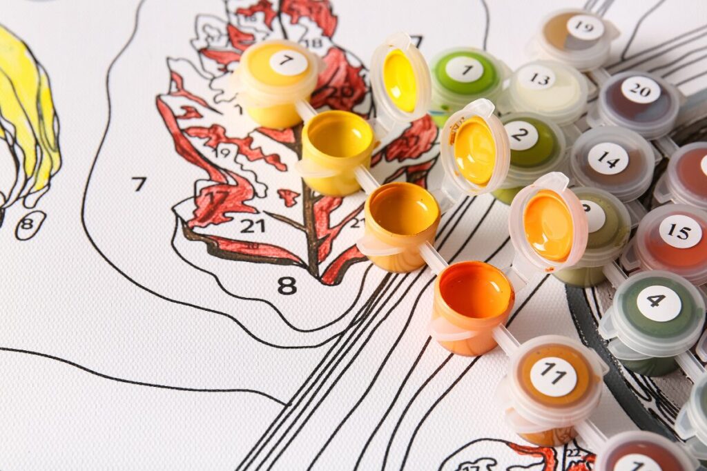 Paint-by-number canvas partially filled with yellow and orange paint next to open paint containers.