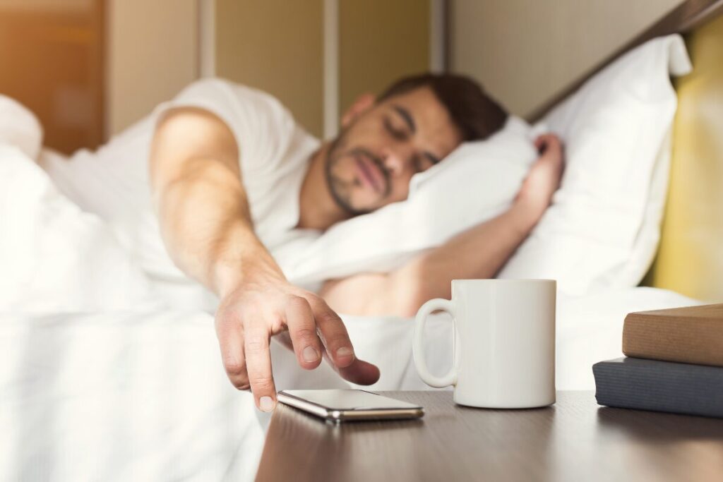 Person reaching for their phone alarm while waking up, showing the reality of starting a morning routine.