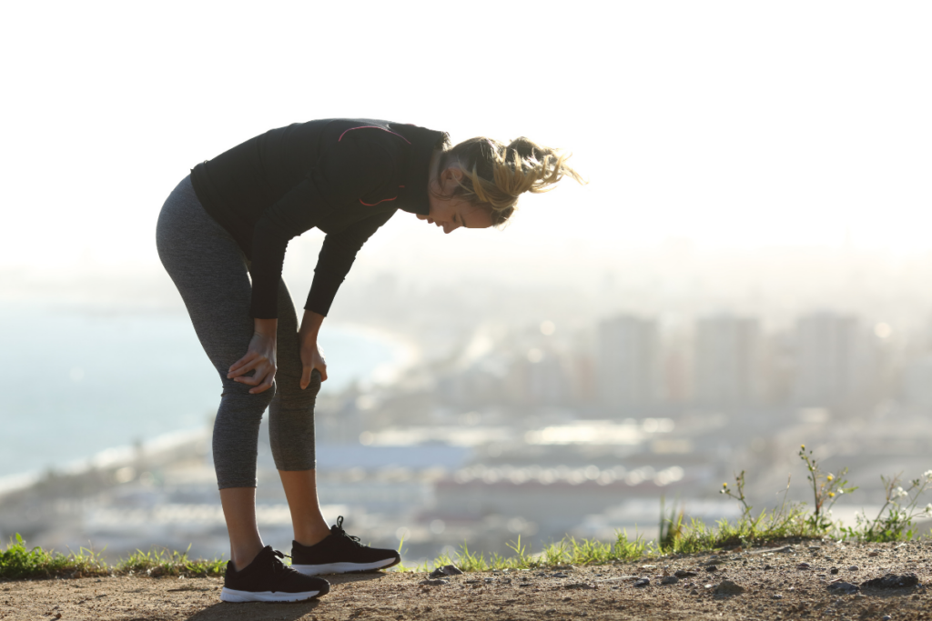 Woman in workout gear bent forward catching her breath on a hilltop overlook after a run