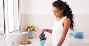 Woman smiling while pouring fresh coffee in a bright kitchen, representing a calm and simple morning routine.
