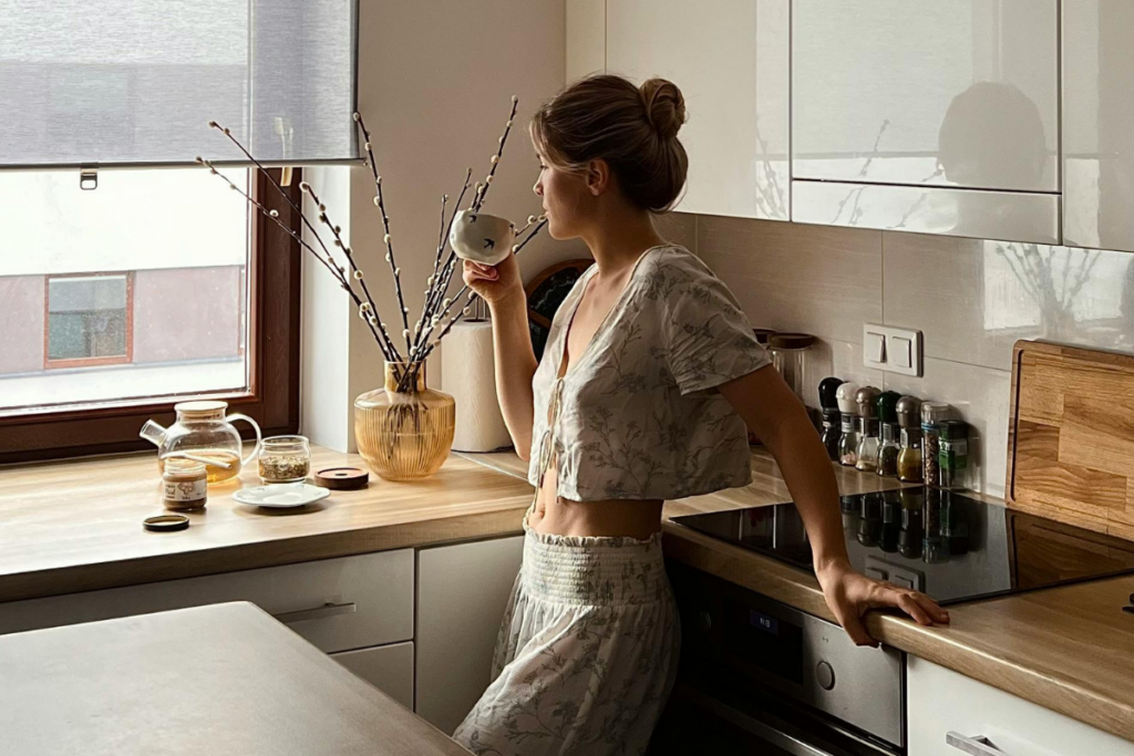 Woman in a floral outfit sipping tea in a sunlit kitchen, gazing out the window in a calm, unhurried morning moment
